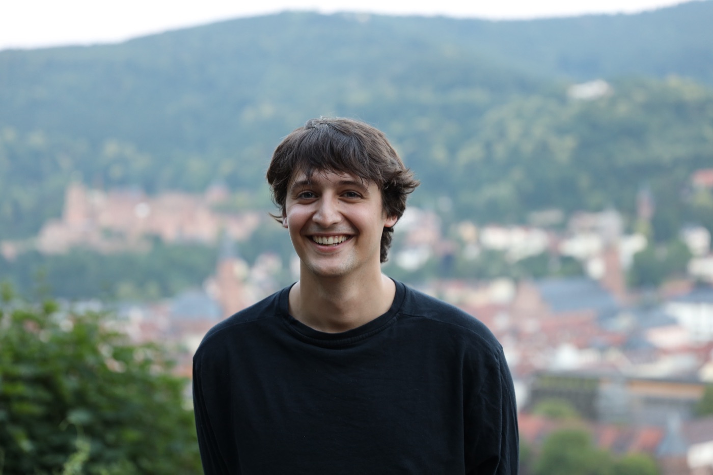 Tiago lächelt, im schwarzen Pullover, hinter ihm unscharf eine Stadt und bewaldete Hügel.